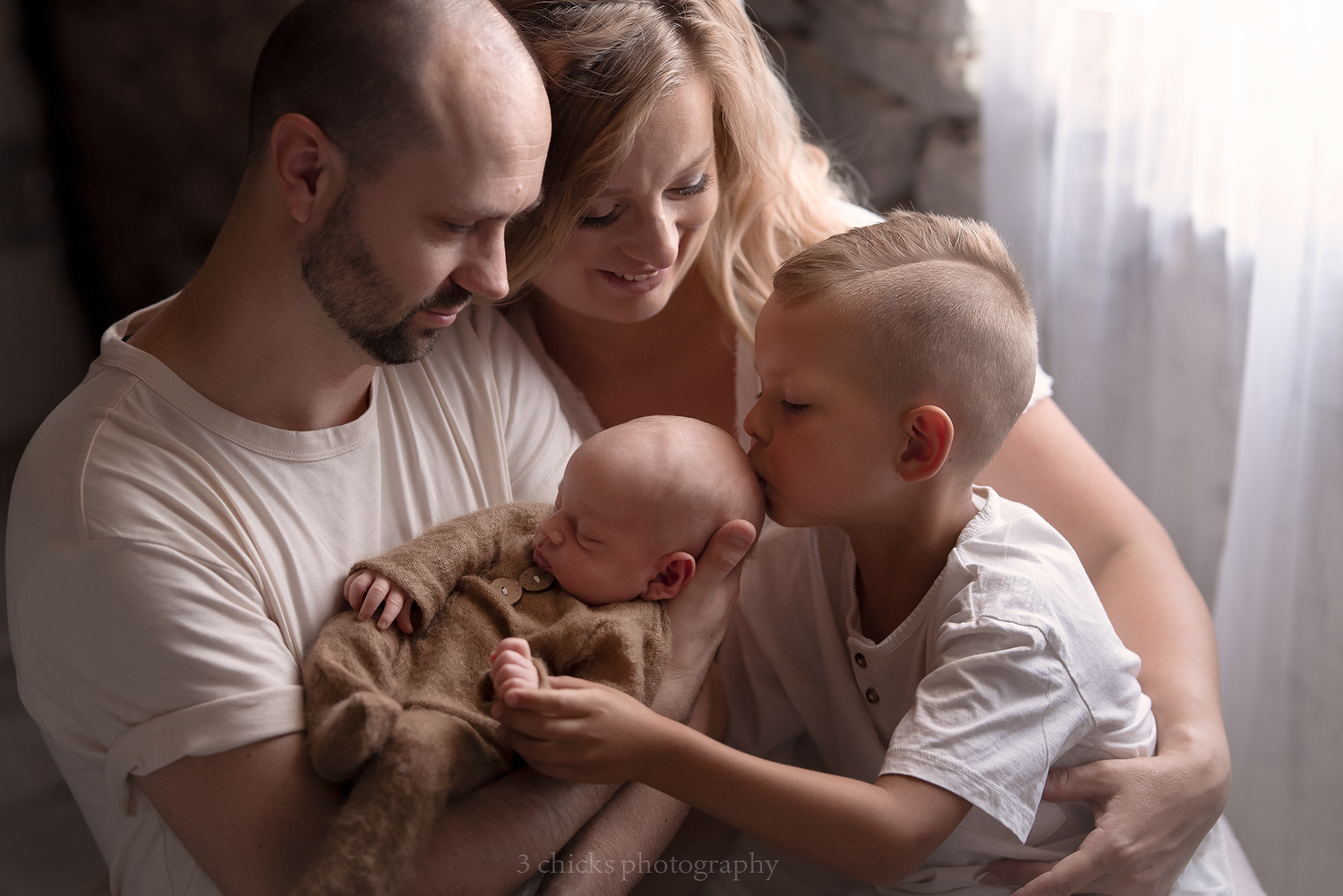 Family of four with parents holding sleeping newborn wrapped in brown, while older brother gives baby a gentle kiss.