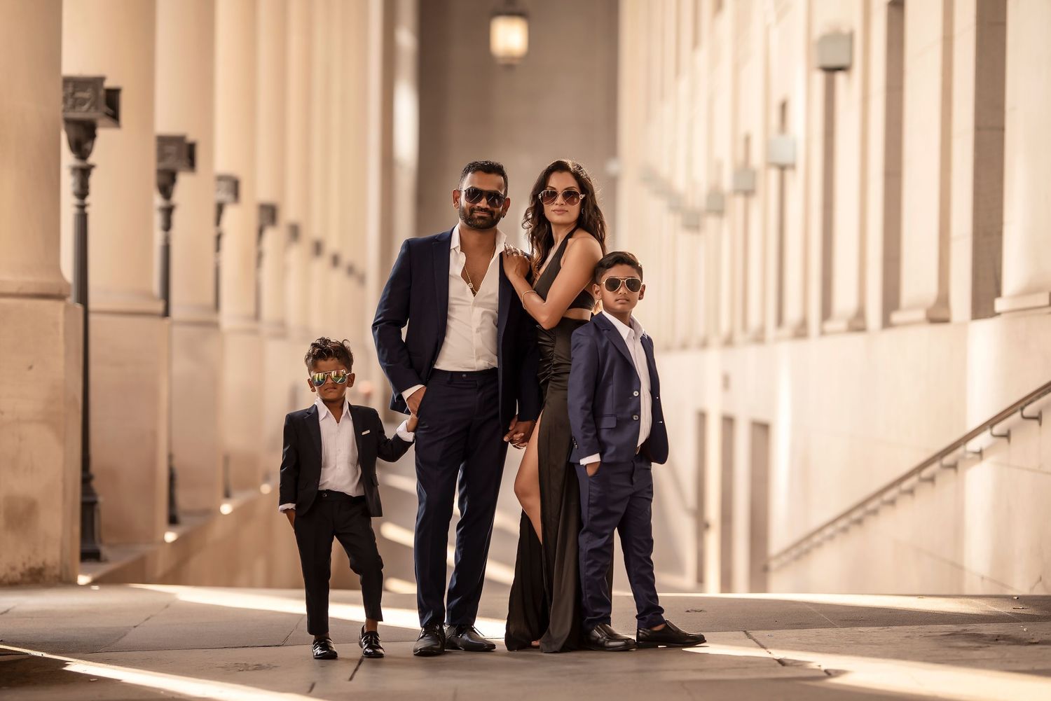 Stylish family of four in elegant dark suits and gown posing in sunlit architectural corridor.