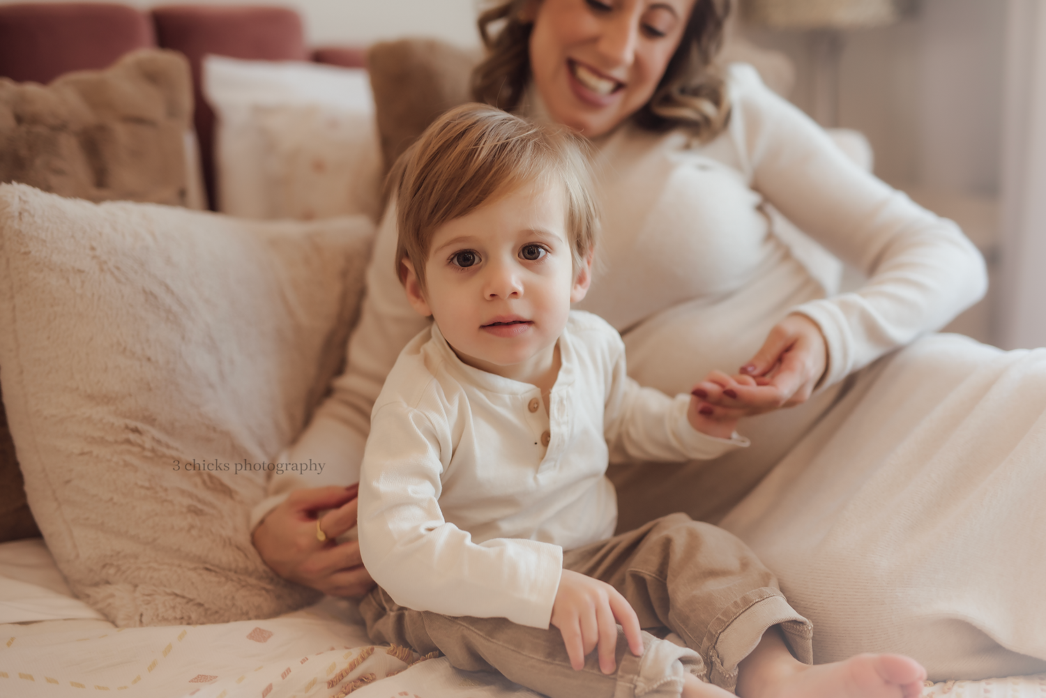 Pregnant mother sitting on a couch smiling while her toddler child sits beside her in a cozy home setting. Baby photography Oakville