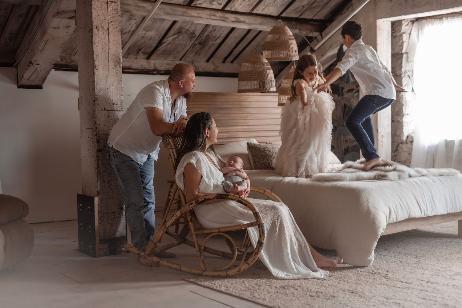 Family enjoys playful moment in rustic bedroom while mother sits in rocking chair holding newborn baby.