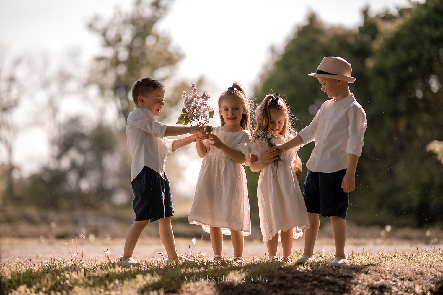 Four children in white outfits sharing a bouquet of purple flowers outdoors in golden sunlight.