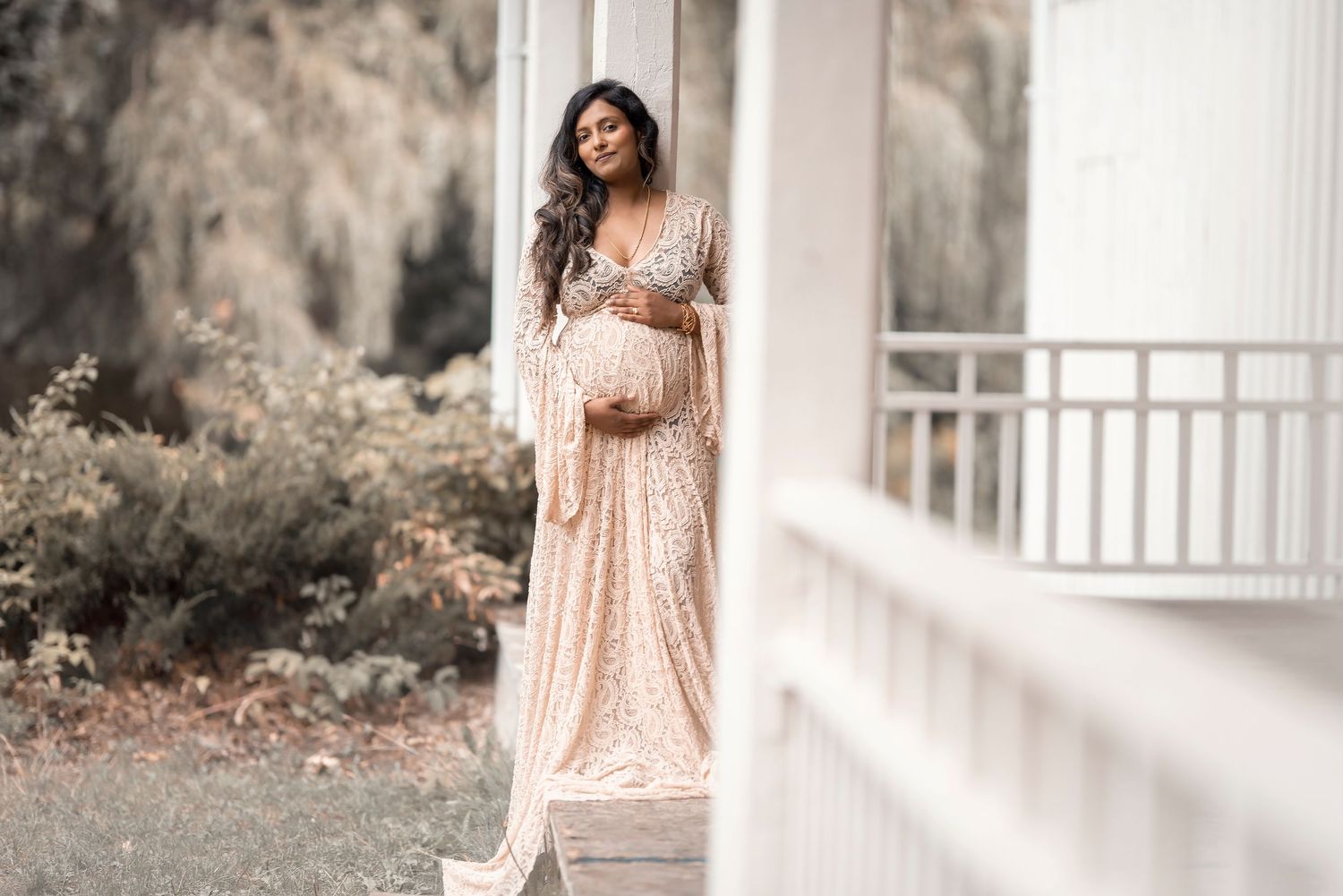 Pregnant woman in sequined blush gown smiling on a white porch outdoors.