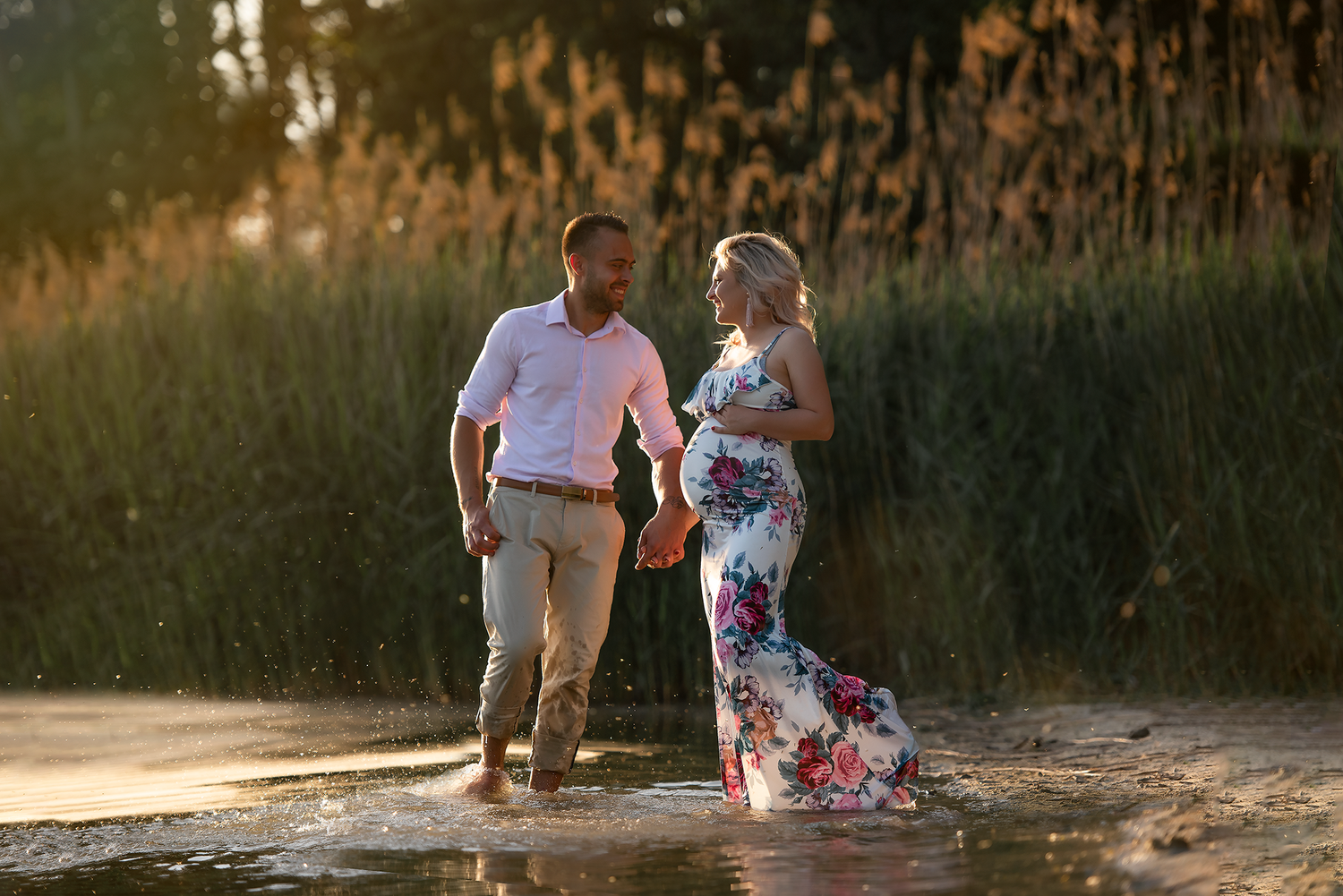 Couple wading through shallow water at golden hour near tall reeds, woman in floral gown, man in white shirt.