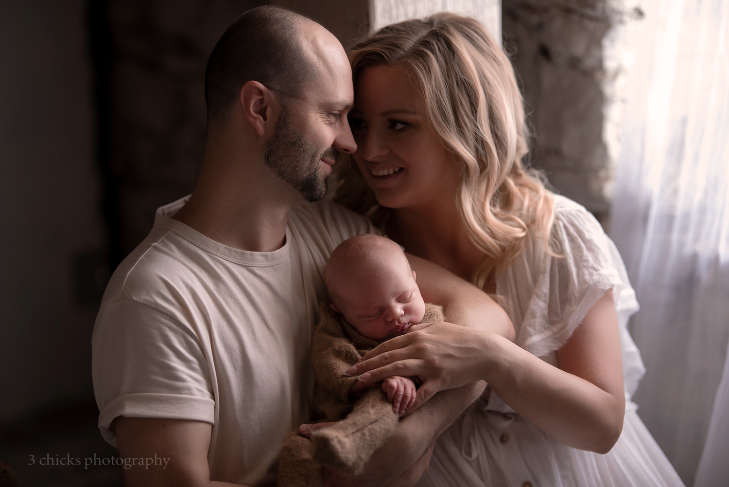 New parents tenderly hold their sleeping newborn baby in a soft, dreamy portrait by J Chicki Photography.