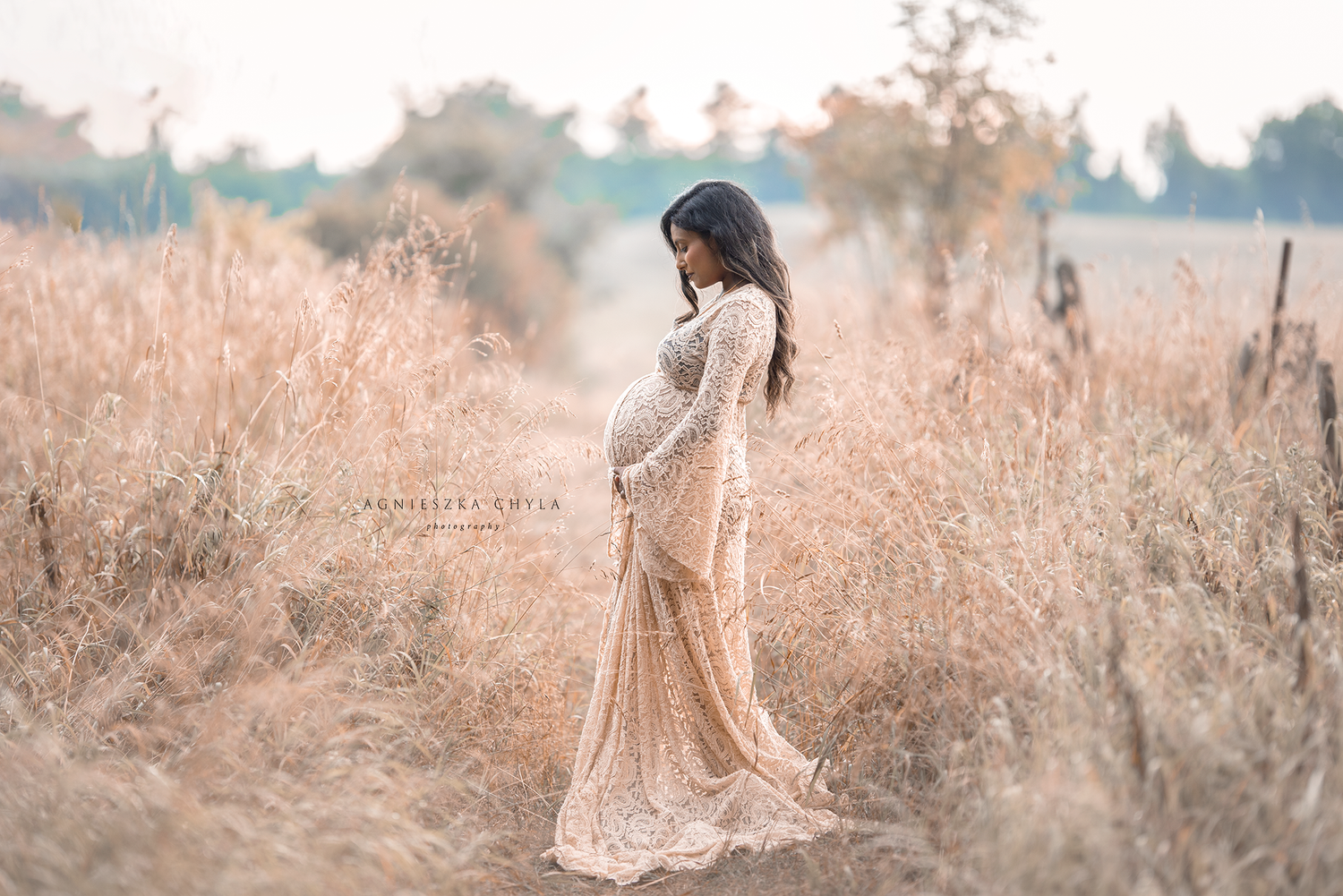 Pregnant woman in lace gown standing in golden field during maternity photo session.