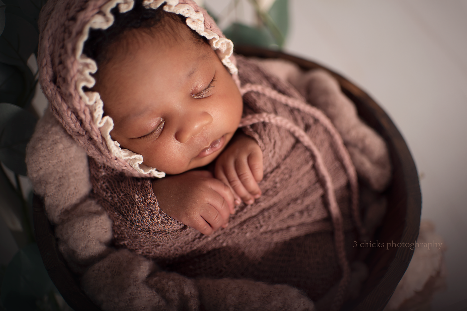 Sleeping newborn baby wrapped in mauve knit blanket wearing a matching bonnet, nestled in a wooden bowl.