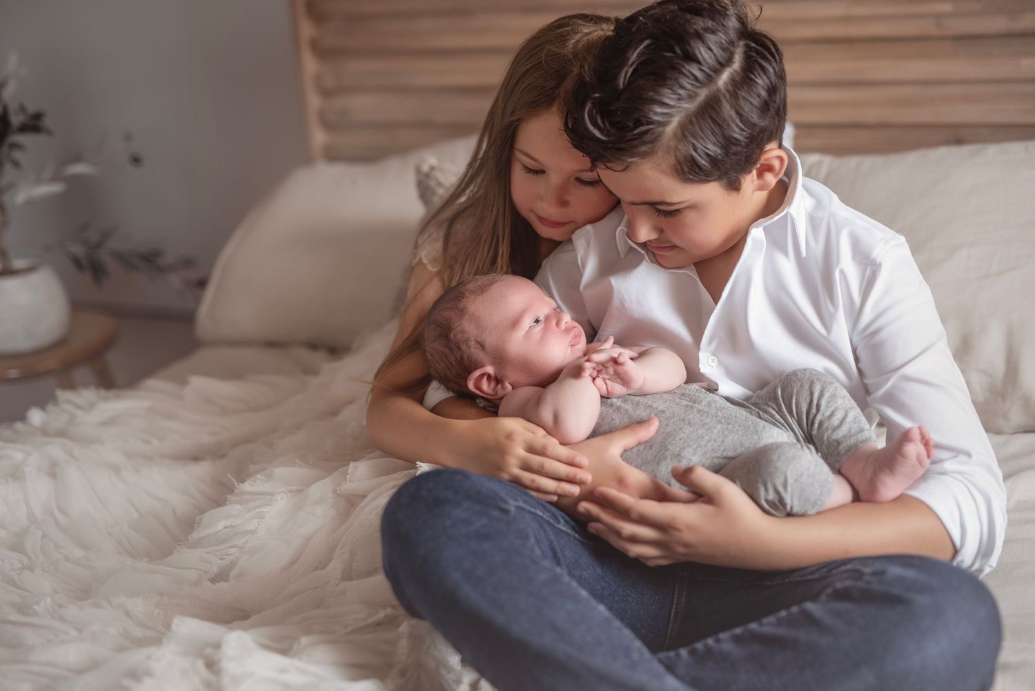 Young girl and mother sit on bed together lovingly cradling a sleeping newborn baby in soft natural light.
