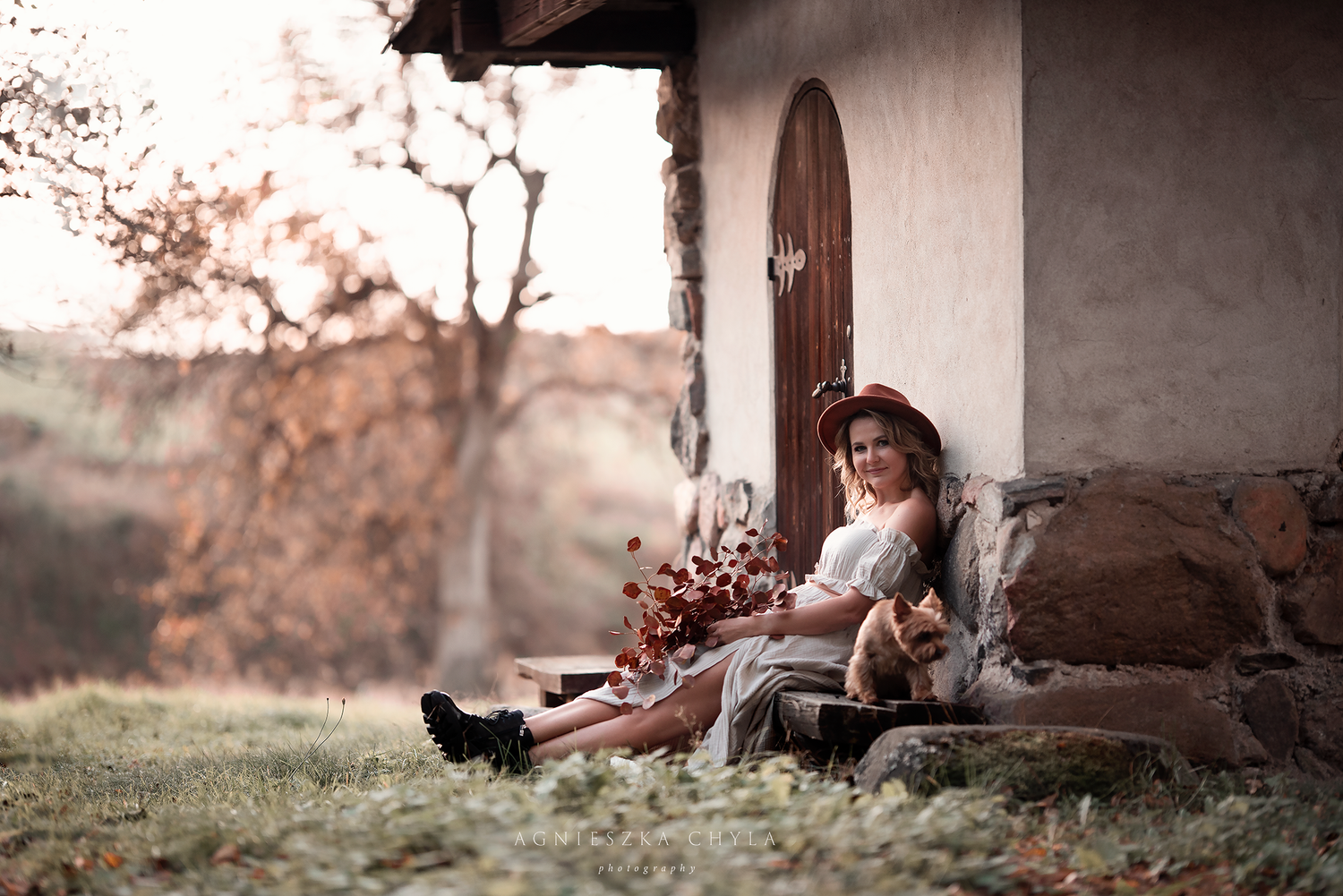 A woman in a hat and midi dress sits against a rustic stone building holding a bouquet of flowers.