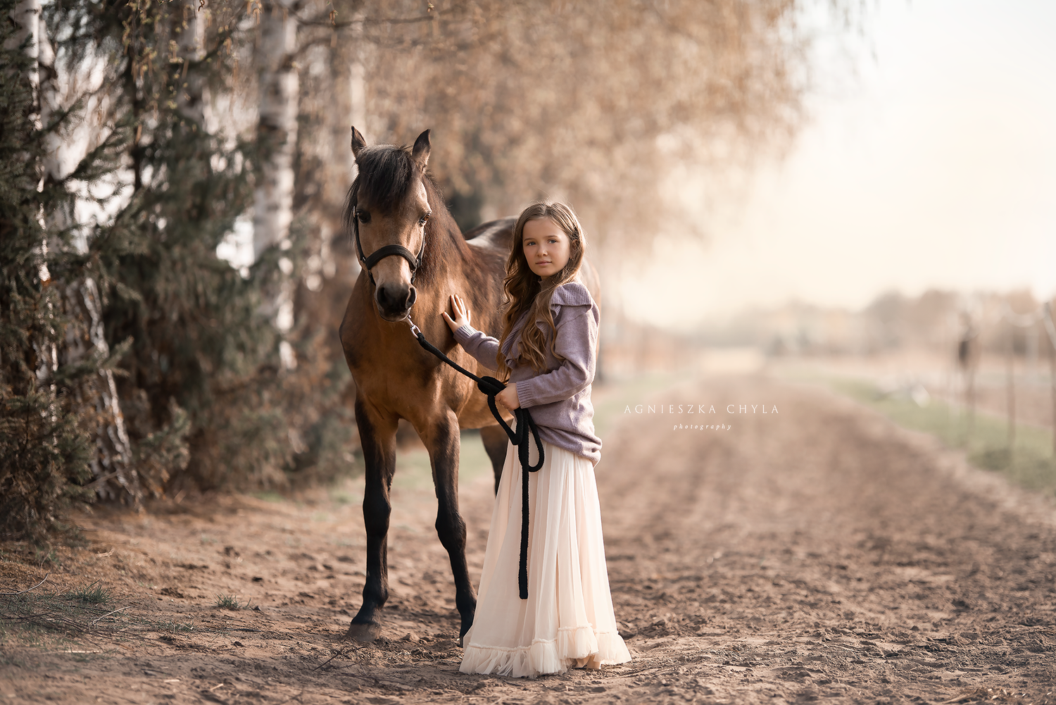 A young girl in a white dress stands beside a bay horse on a sandy path surrounded by autumn trees.
