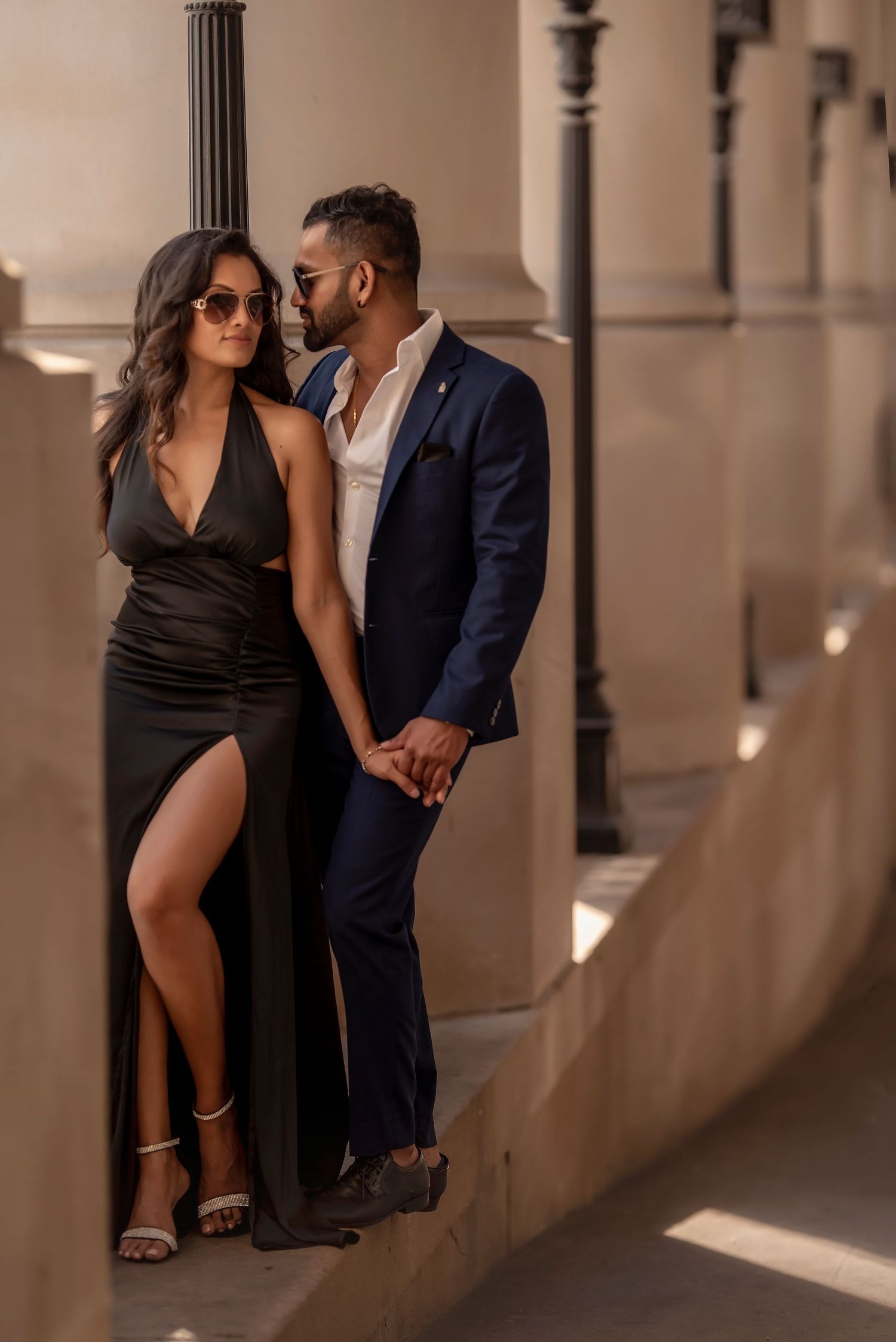 Elegant couple in formal attire posing between columns in warm sunlight.