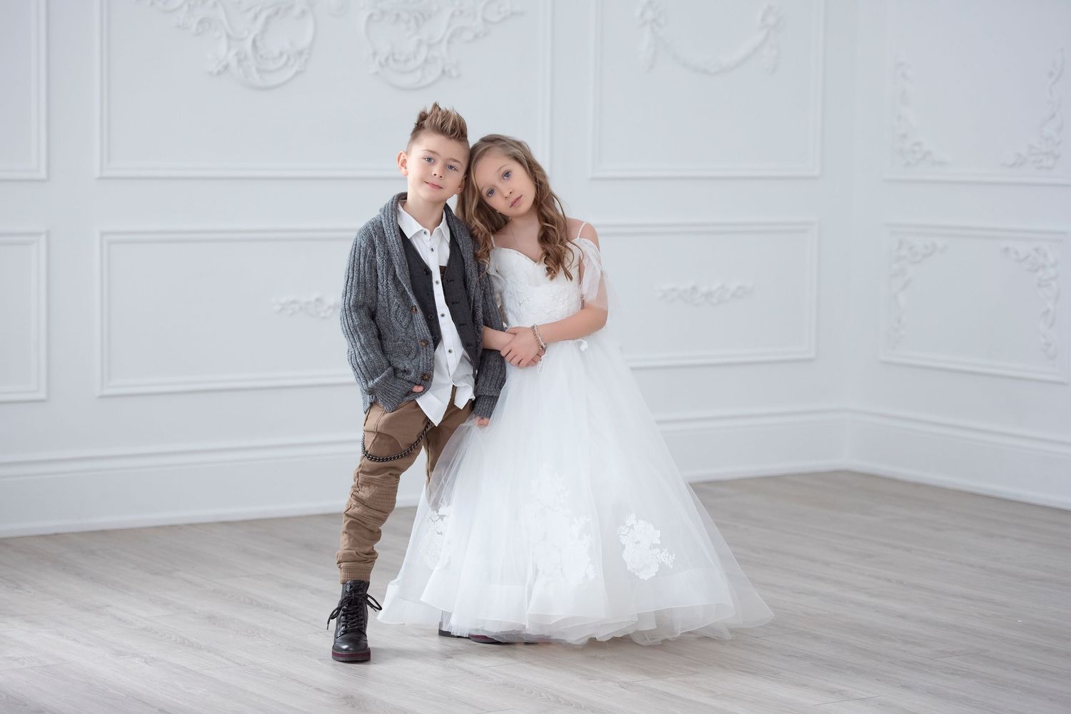 A boy in casual attire and a girl in a white ball gown pose together in a white room. Kids photography studio session Burlington