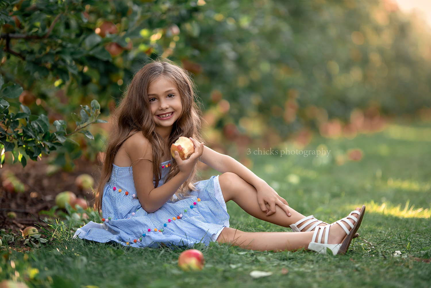 Smiling girl in blue dress sitting on grass holding an apple in orchard.
