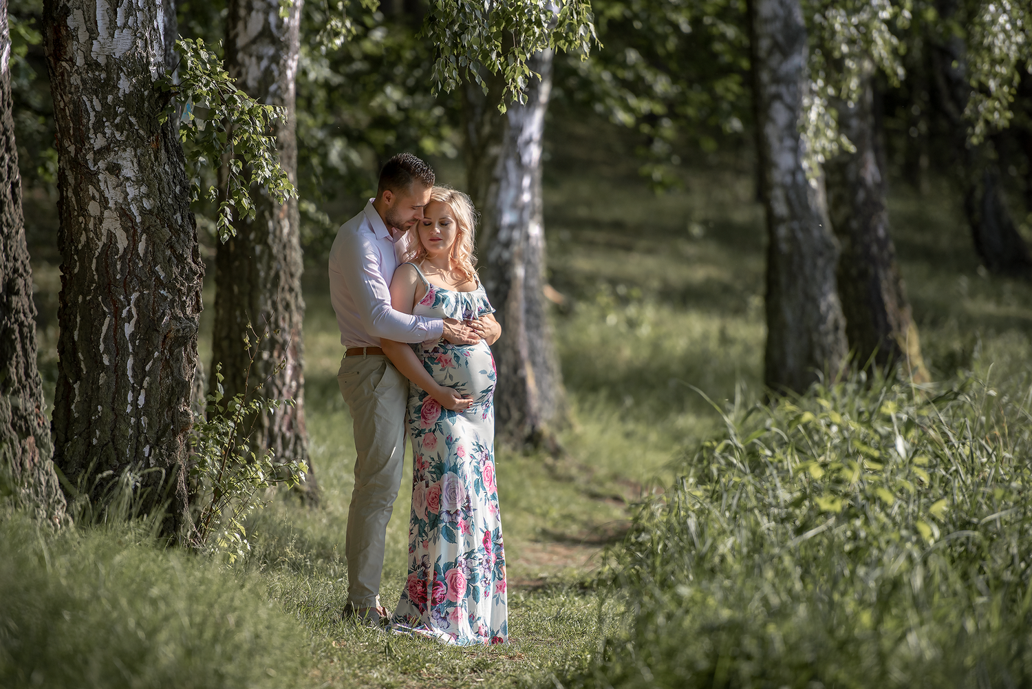 A couple embraces tenderly in a birch forest, the woman in a floral dress showing a pregnant belly.