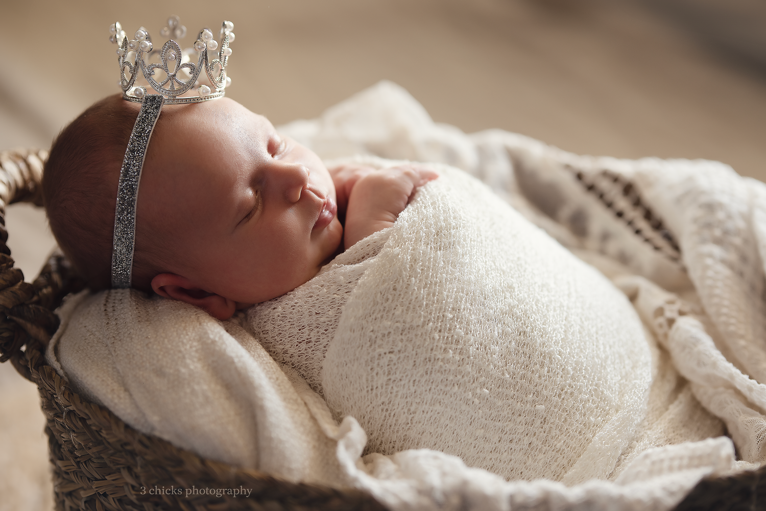 Newborn baby swaddled in white wrap wearing a tiny silver crown, sleeping peacefully in a rustic basket.