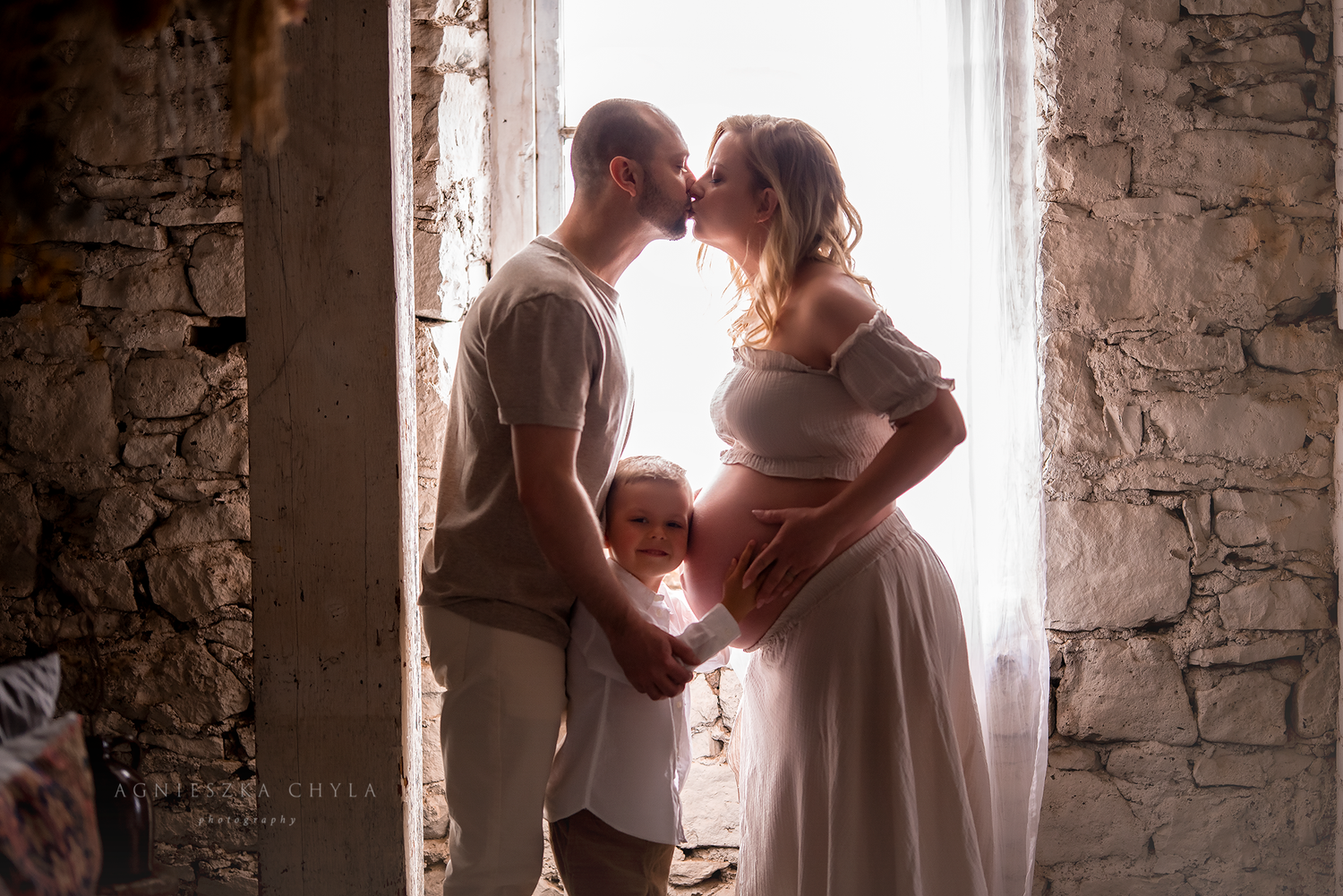 Couple sharing a tender kiss in soft backlit window light during a maternity photo session with toddler.