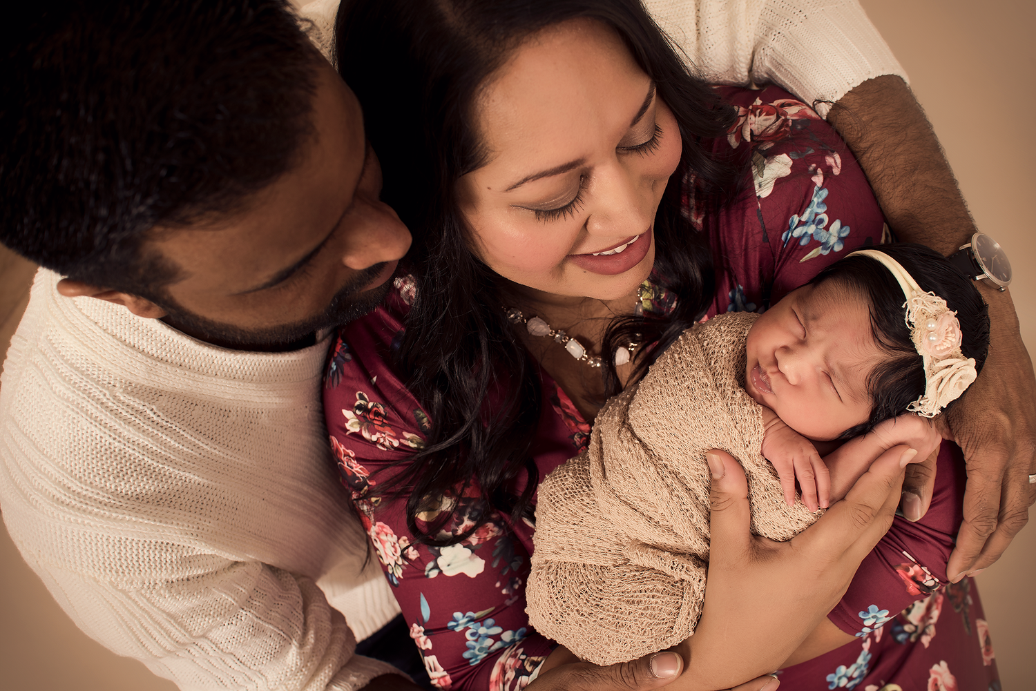 New parents lovingly gaze at their sleeping newborn baby wrapped in a cream knit blanket with a floral headband.