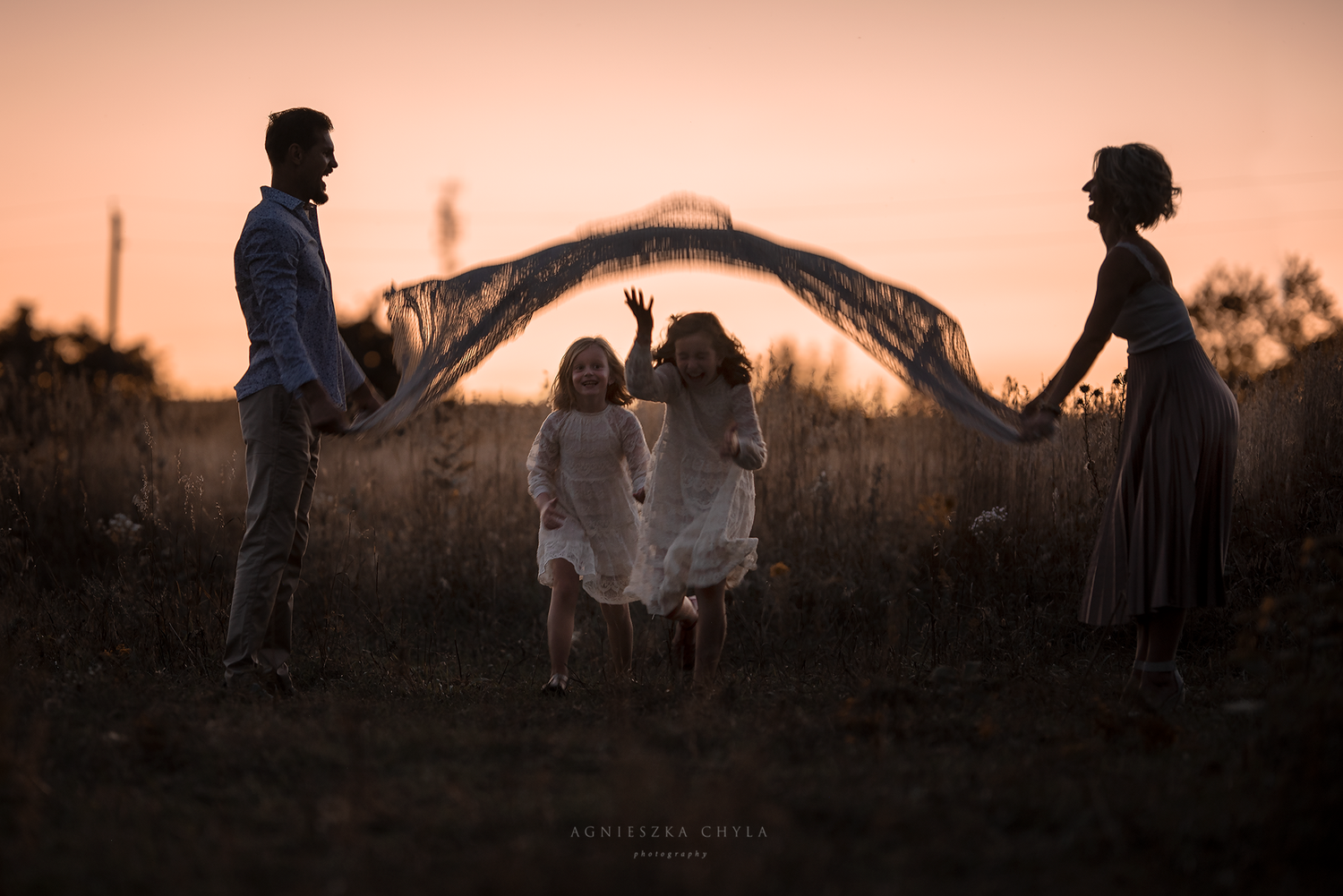 Silhouette of family with two children playing under flowing fabric at golden sunset in open field.