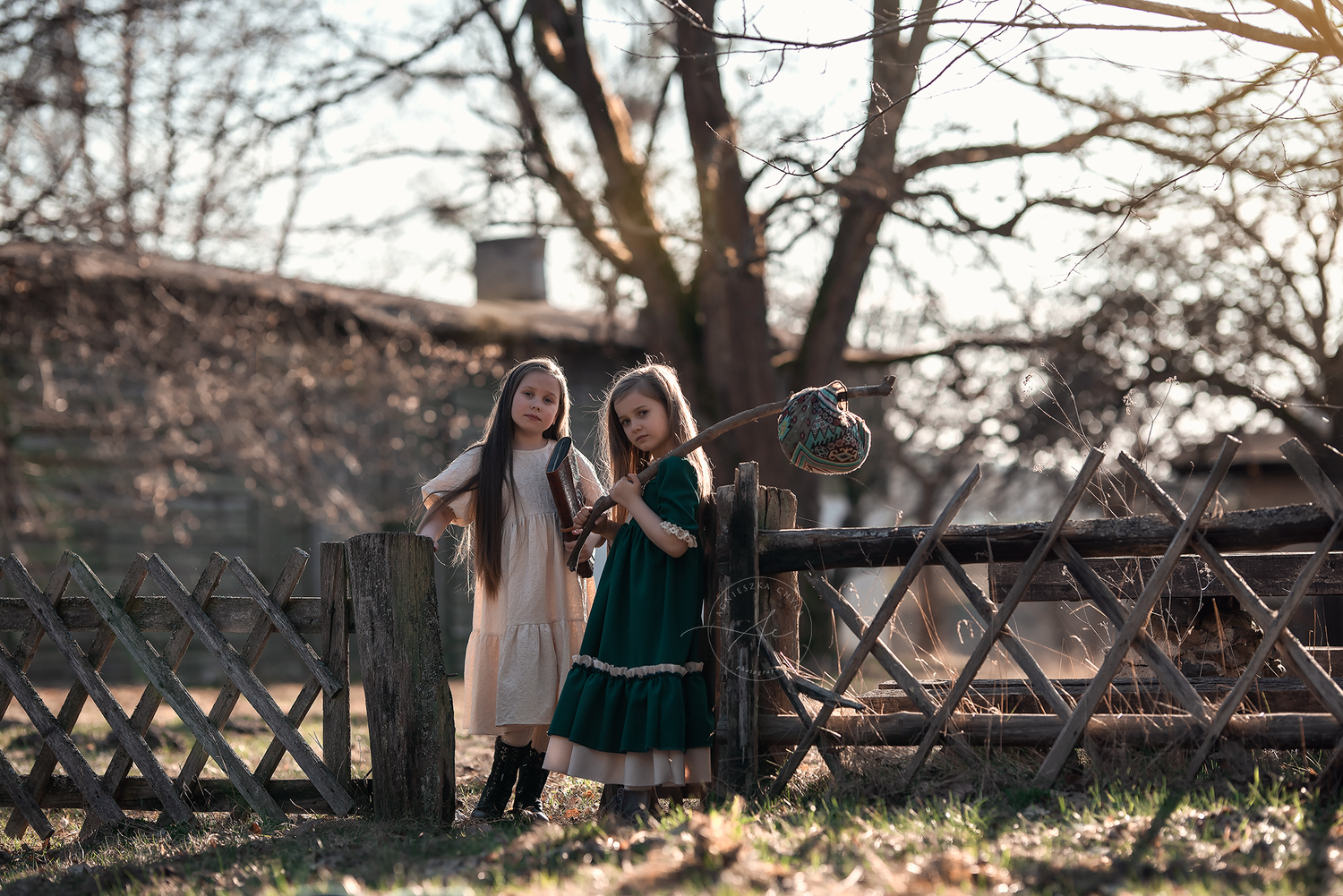 Two girls in vintage dresses standing by a rustic wooden fence in autumn countryside setting.