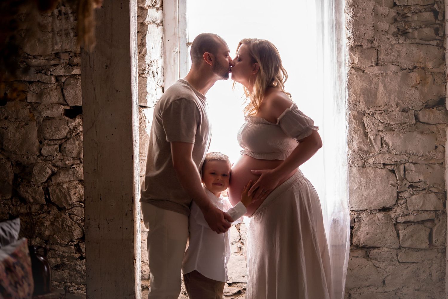 Couple kissing with toddler between them in a sunlit stone archway during maternity shoot.