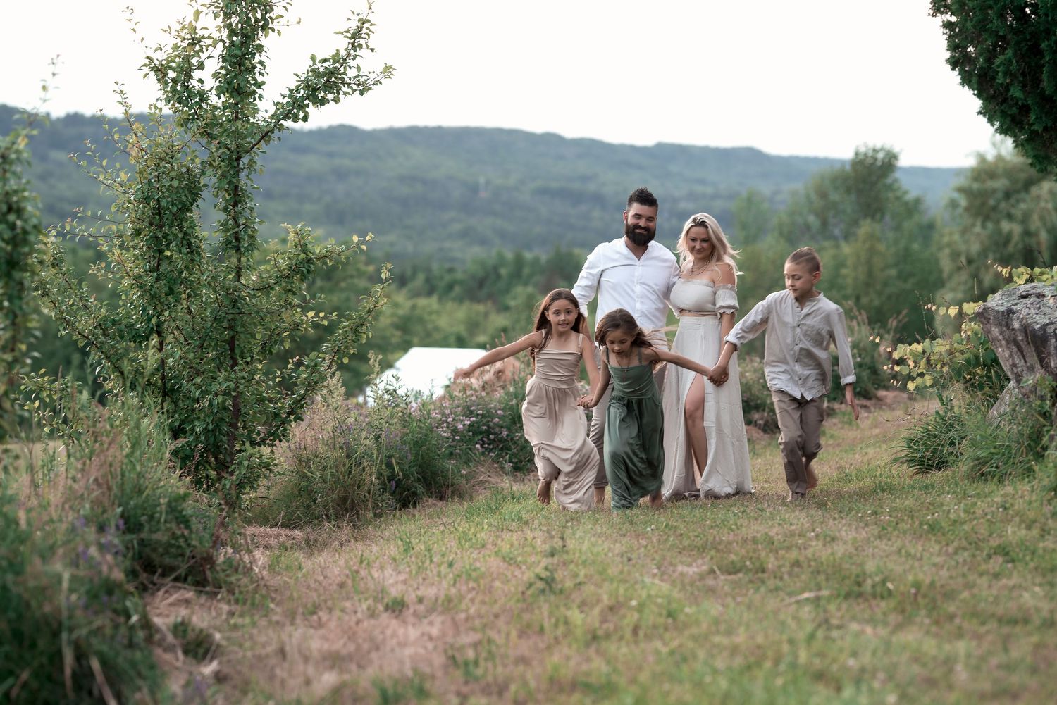 A joyful family with children walks hand-in-hand through a lush green hillside landscape near a lake.