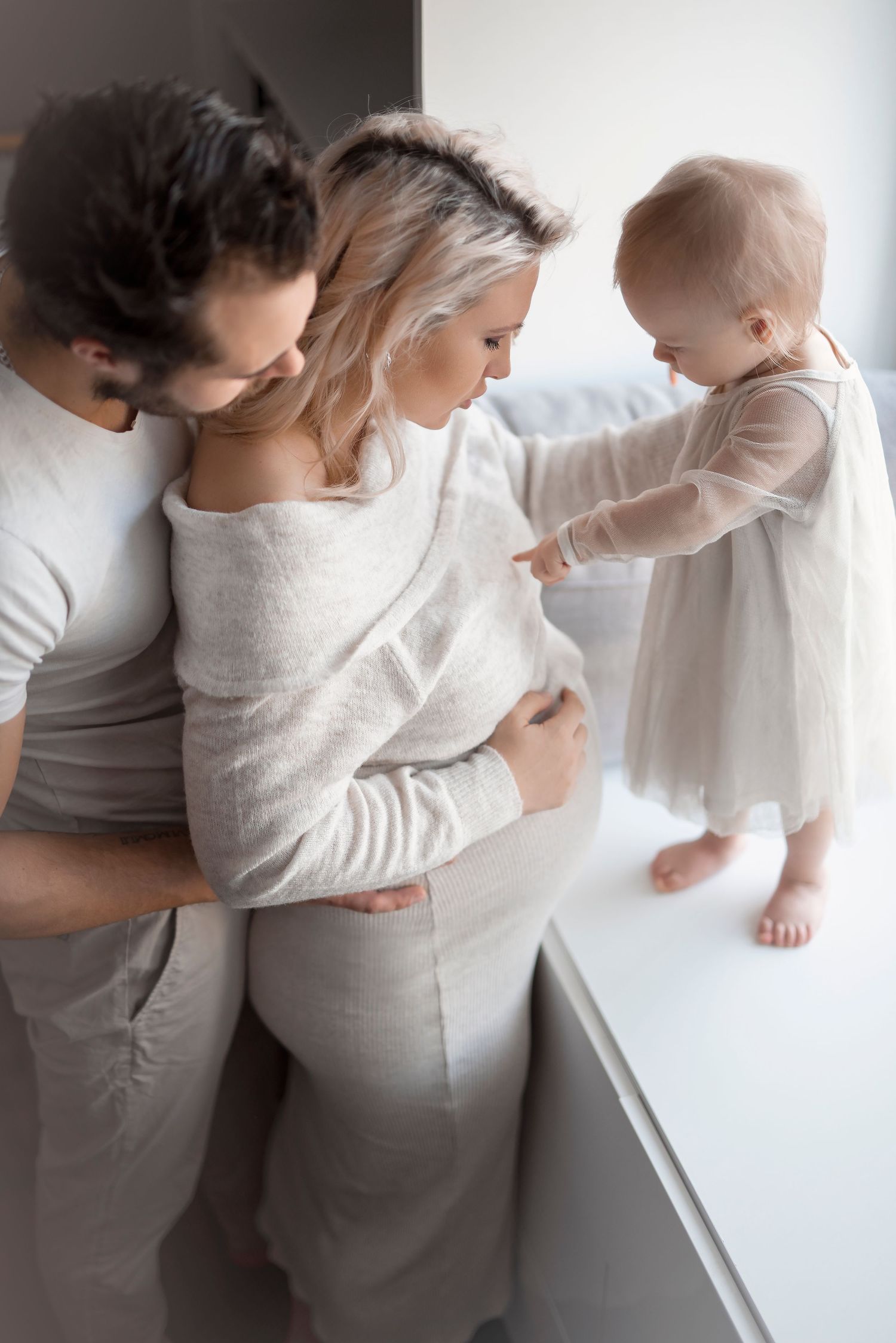 Pregnant woman in white linen dress with husband and toddler daughter touching baby bump at home.
