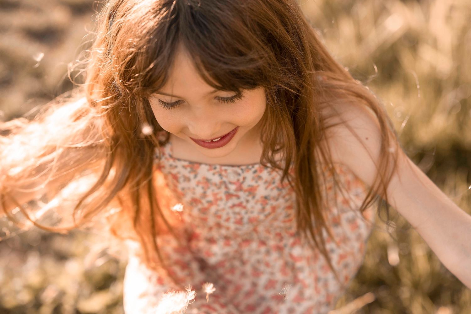 Young girl with long brown hair smiling down at wildflowers in a sunlit field wearing a floral dress.