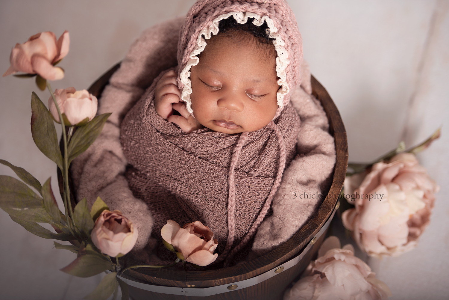 Newborn baby sleeping peacefully in a rustic bucket, wrapped in mauve knit, wearing a lace bonnet, surrounded by pink roses.