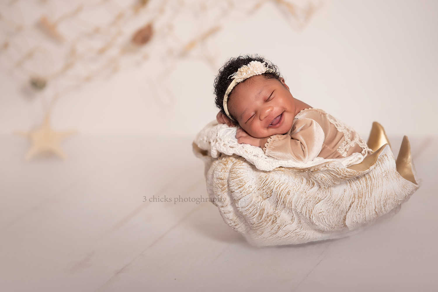 Smiling newborn baby in cream lace outfit and tiara rests in decorative moon prop with gold stars backdrop.