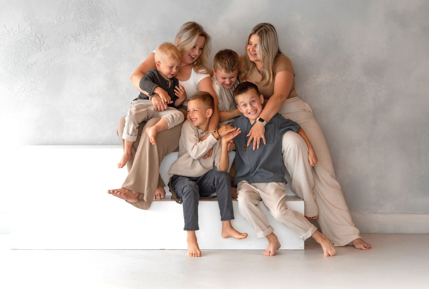 Happy family of five posing together on white steps against a gray wall in a warm, casual studio portrait.