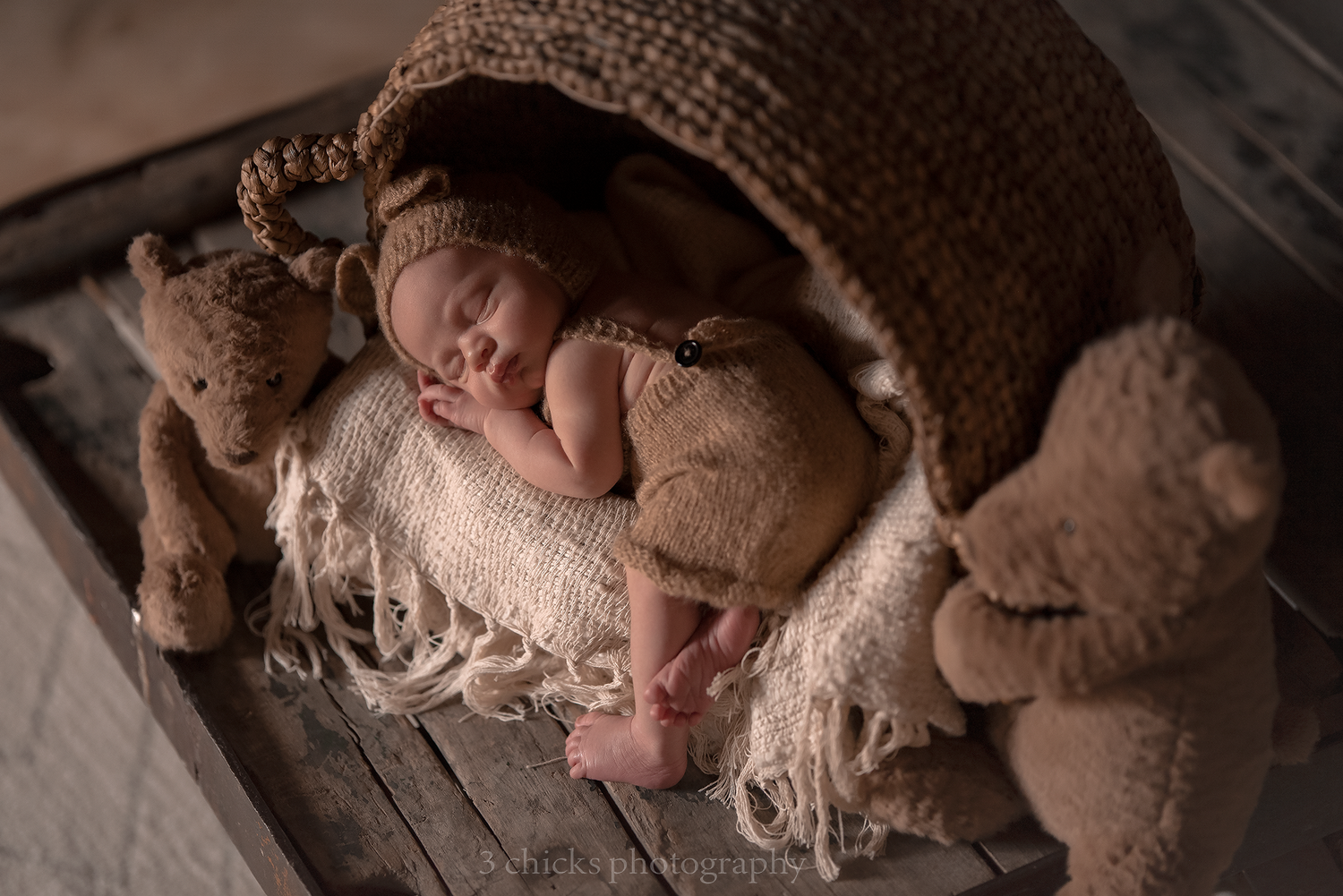 Sleeping newborn baby in brown knit bonnet nestled between two teddy bears in a rustic wooden crate.