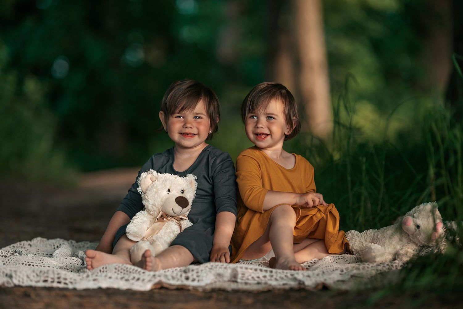 Two smiling toddlers sitting on a blanket outdoors with a white teddy bear.