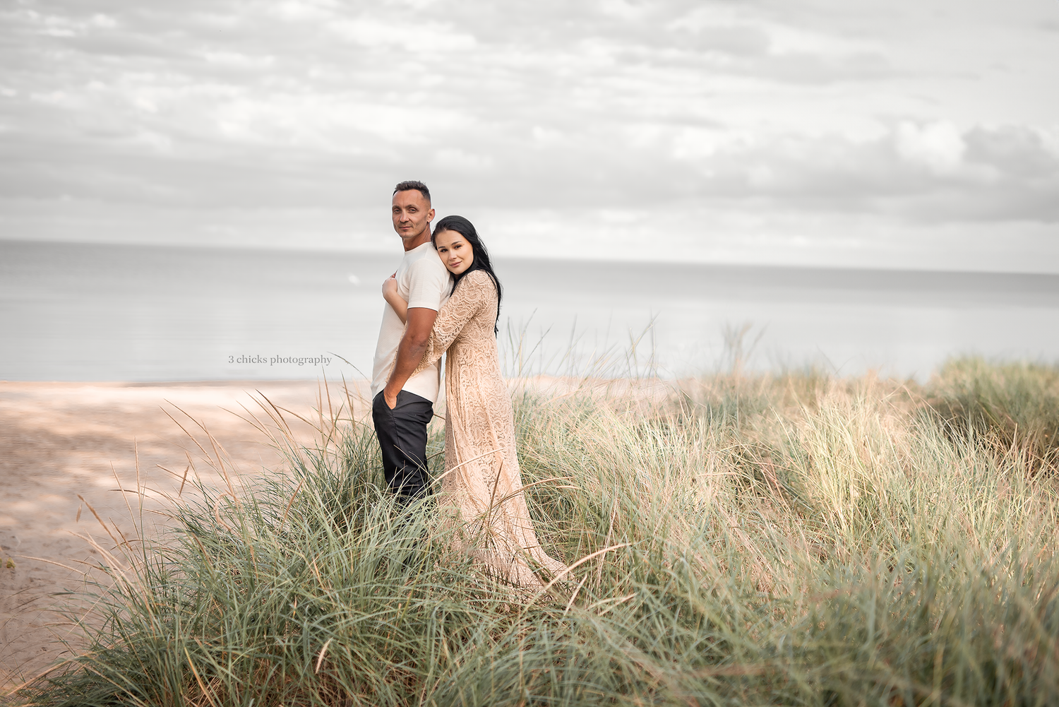 Couple standing close together in coastal dune grass with calm ocean in the background.