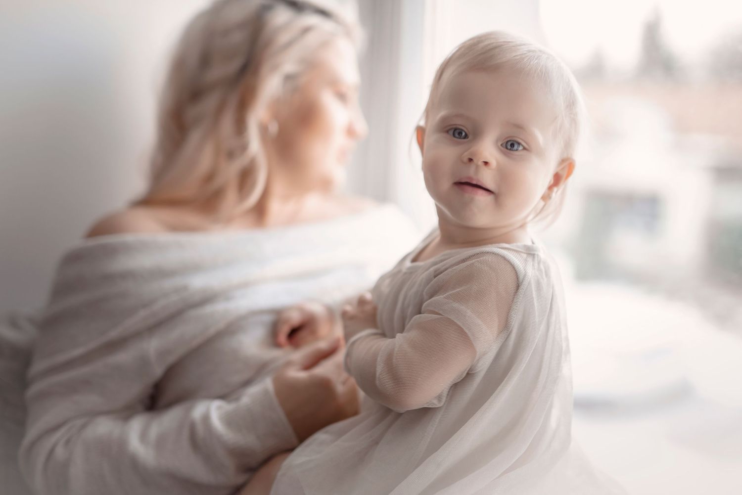 A blonde mother holds her smiling baby in a white dress near a bright window in a soft, dreamy portrait.