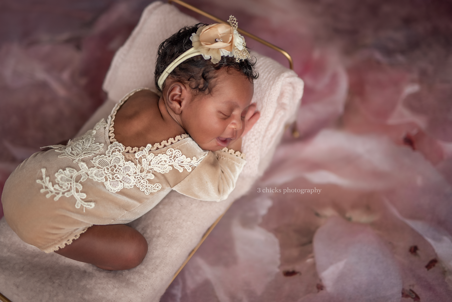 Newborn baby girl sleeping peacefully in lace romper and floral headband on pink tulle backdrop.