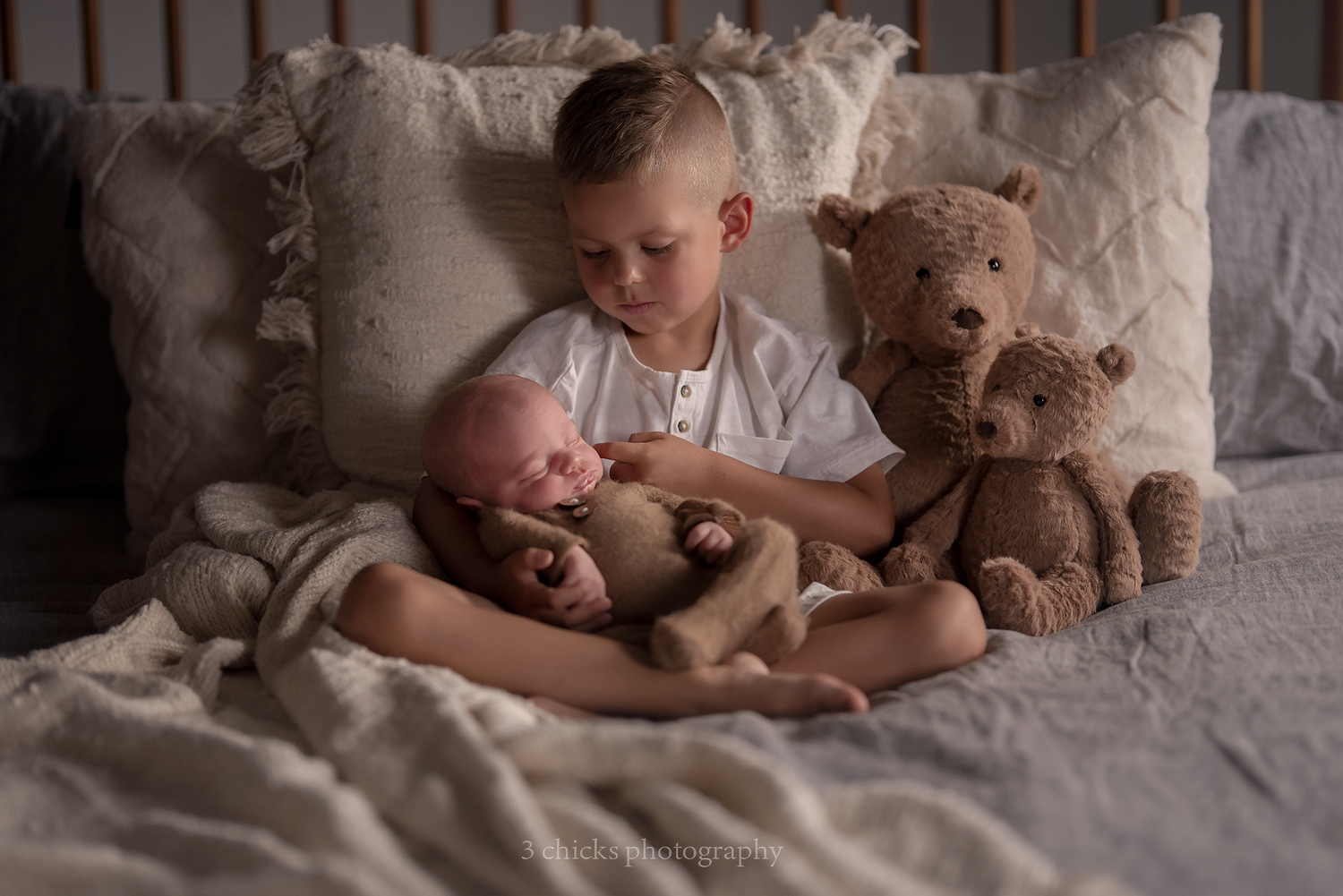 Young boy sitting on bed holding sleeping newborn sibling with teddy bears.