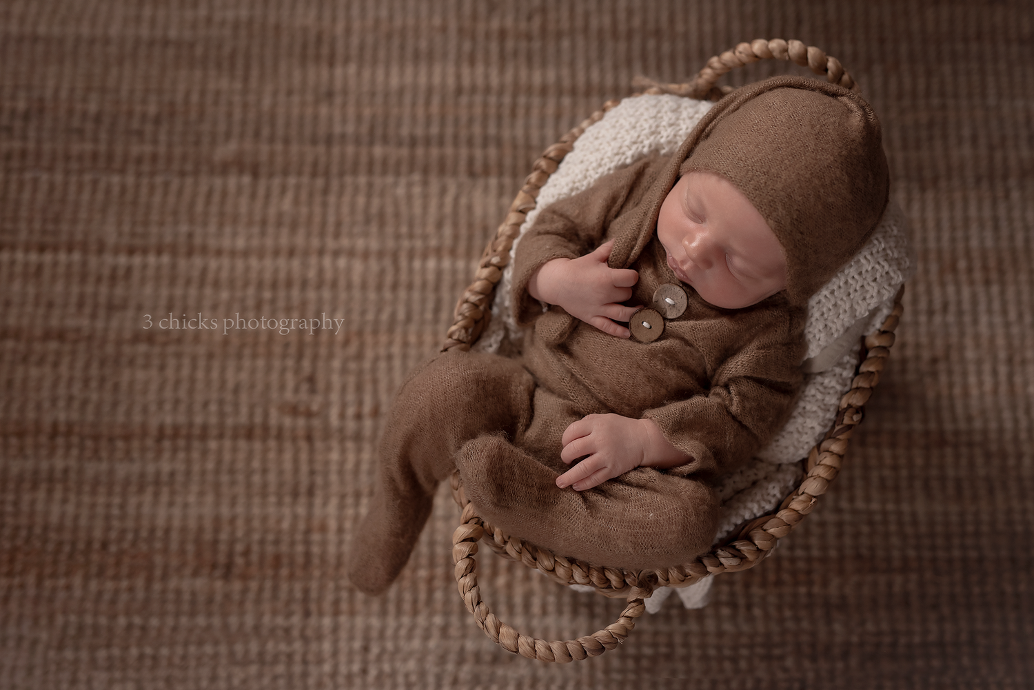 Newborn baby sleeping peacefully in a round wicker basket, dressed in a brown knit romper and matching bonnet hat.