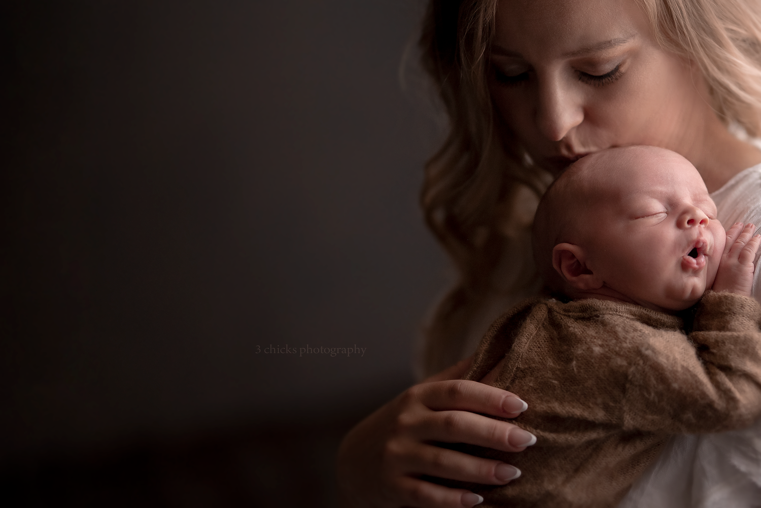 Mother tenderly kissing her sleeping newborn baby wrapped in soft brown blanket.