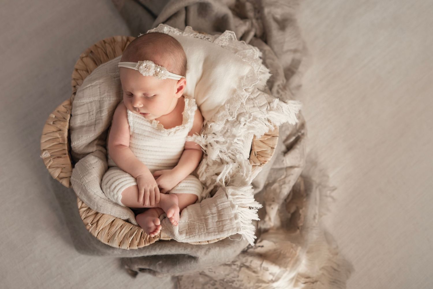 Peaceful newborn baby sleeping in a wicker basket with soft cream blankets and a delicate floral headband.