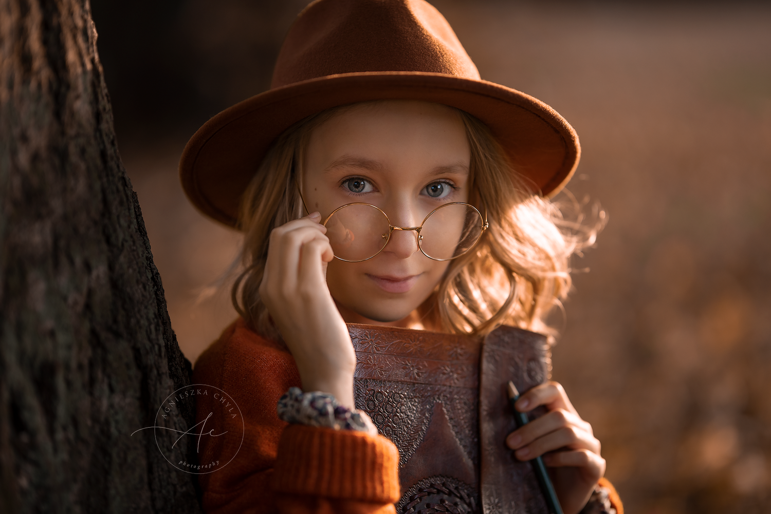 Young girl in orange sweater and brown hat posing by tree in warm light. Headshots and portraits Oakville