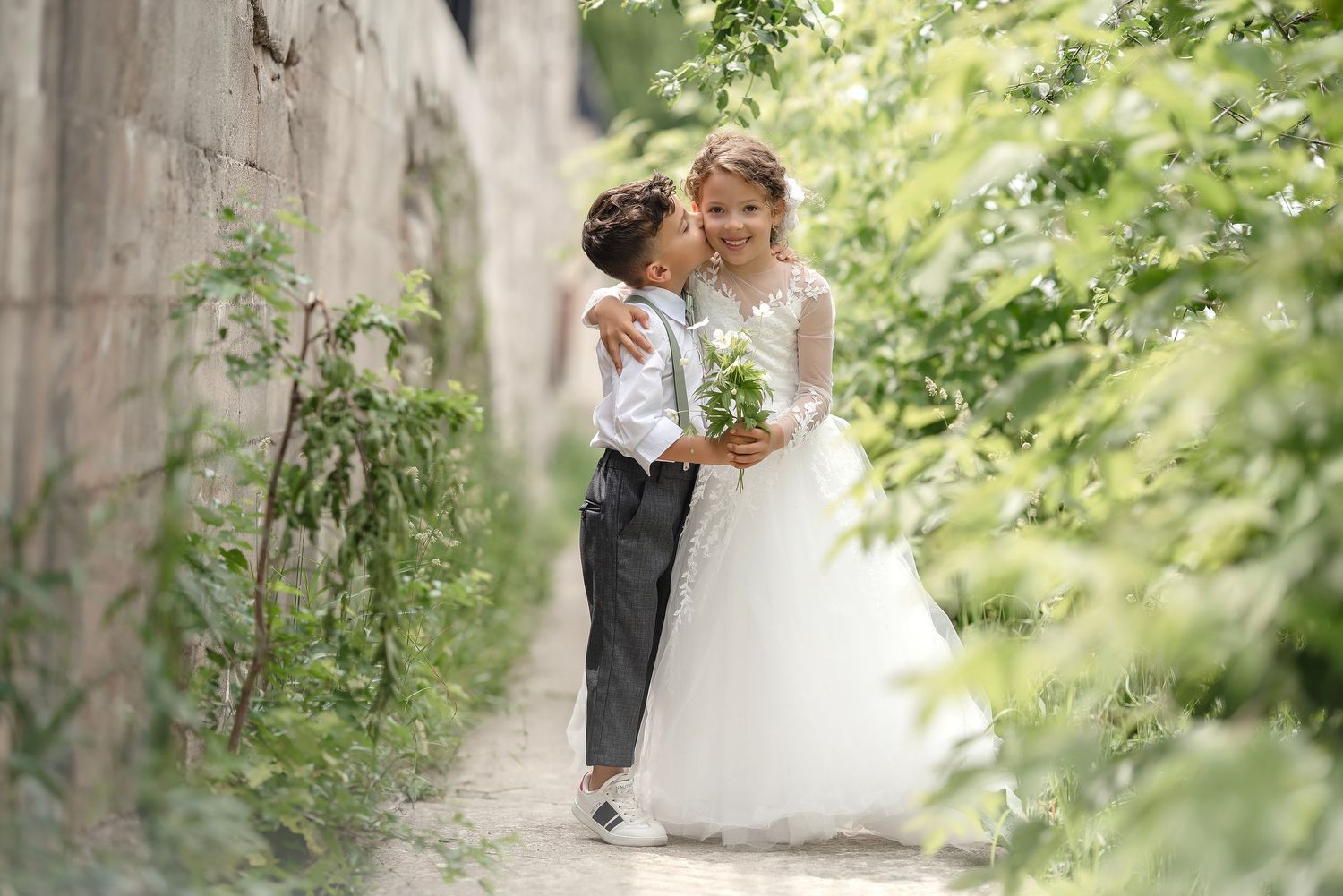 Children dressed as bride and groom embracing in garden pathway.