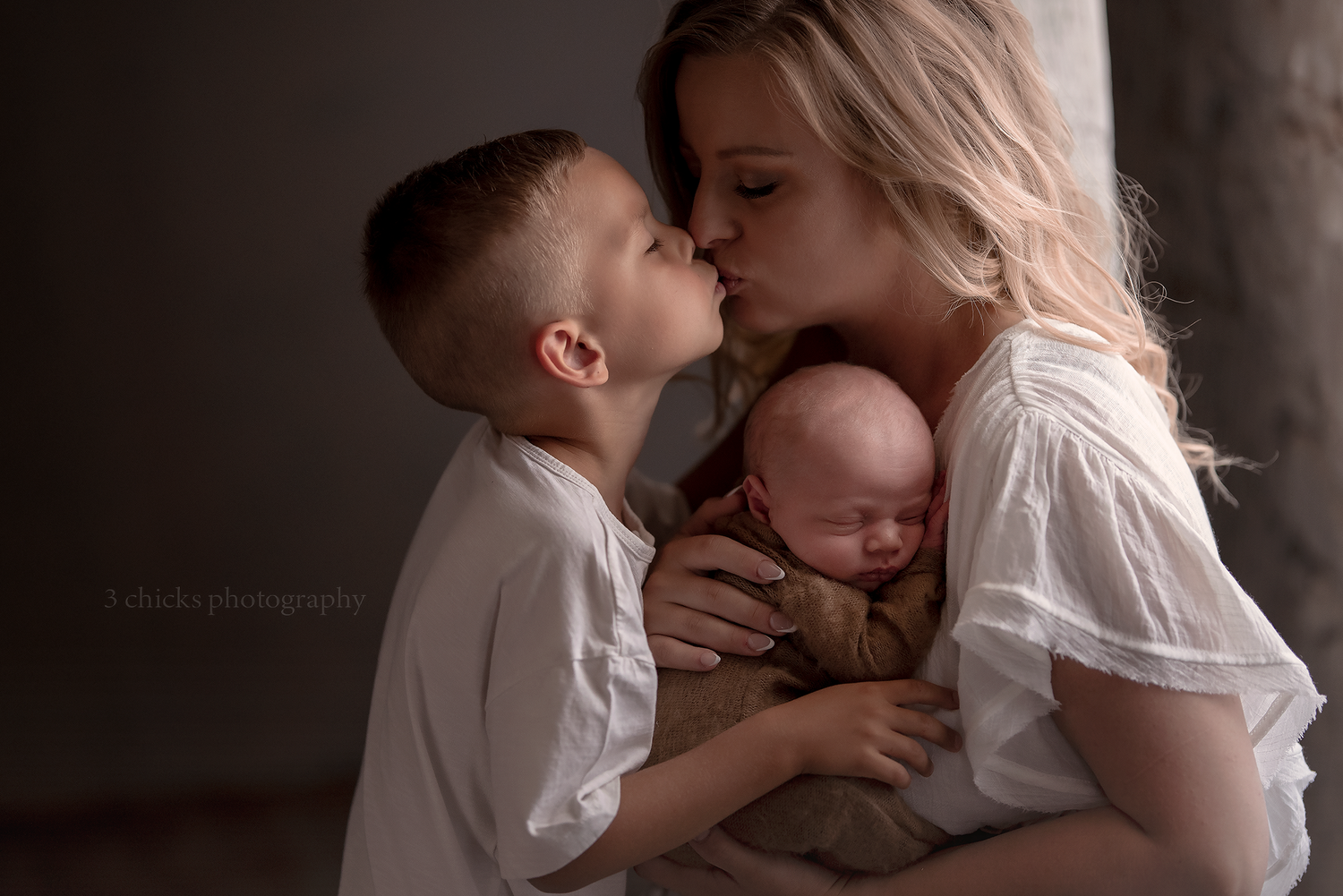 A mother in white dress holds newborn while young boy gives her a tender kiss in soft studio lighting.