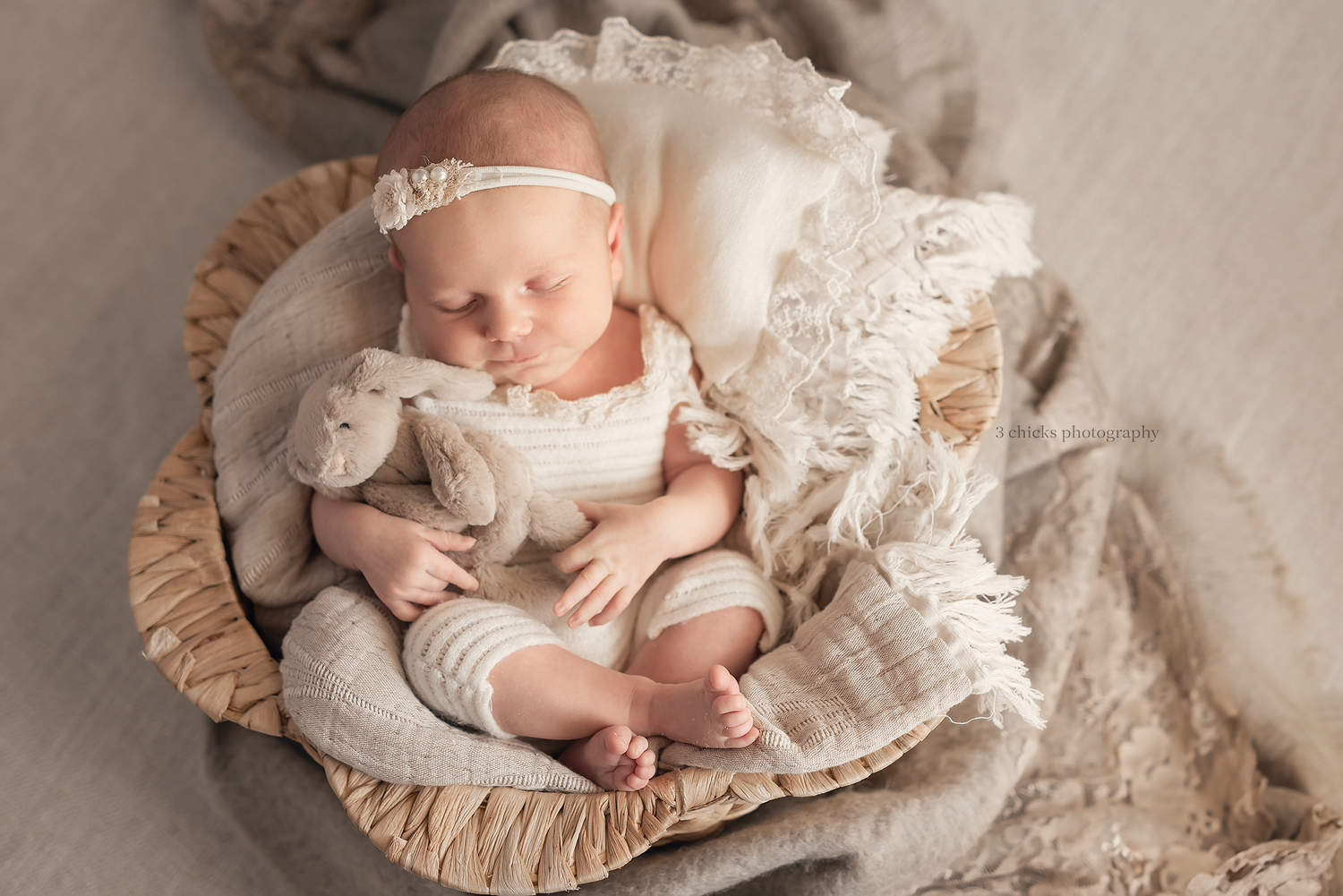 Sleeping newborn baby in a wicker basket with a stuffed bunny, dressed in cream romper and lace headband.