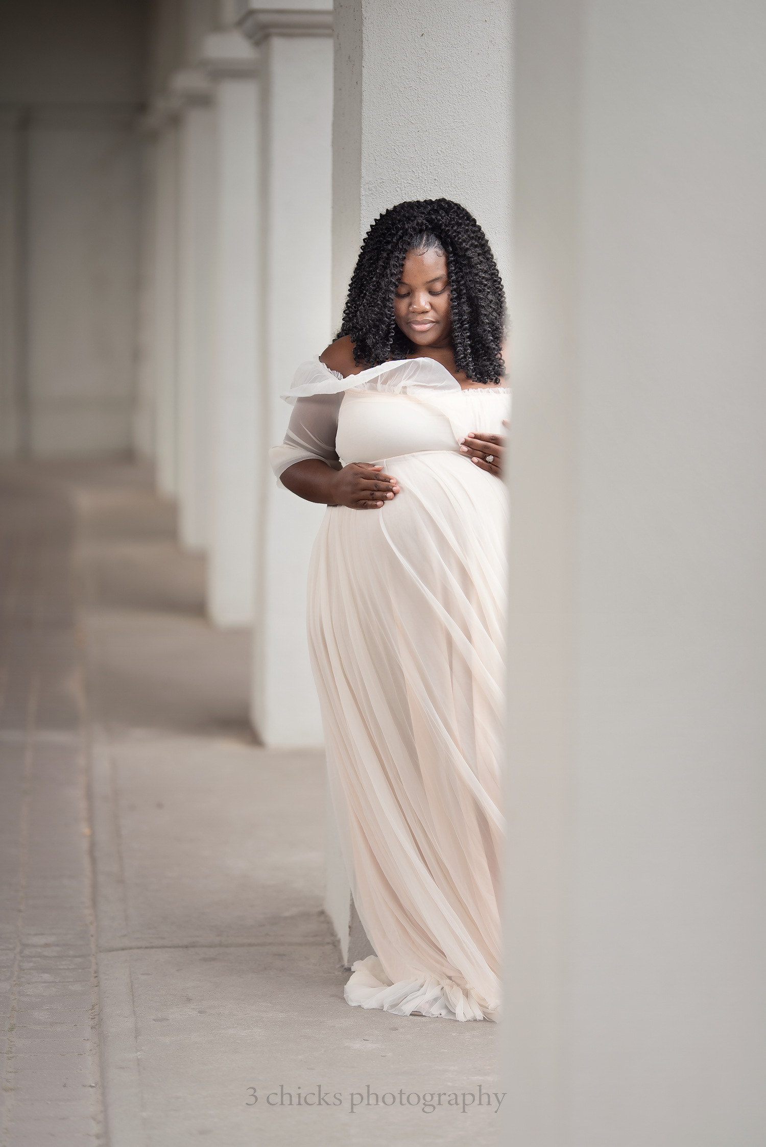 Pregnant woman in elegant off-shoulder cream gown cradling baby bump between white columns, maternity photography.