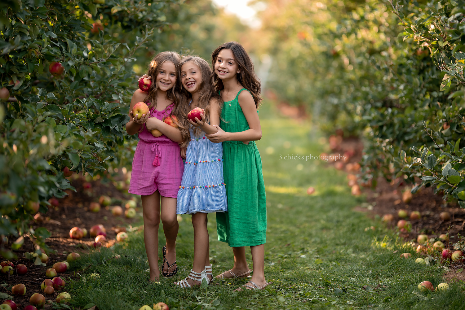 Three smiling girls in colorful dresses holding apples while standing in a lush apple orchard.