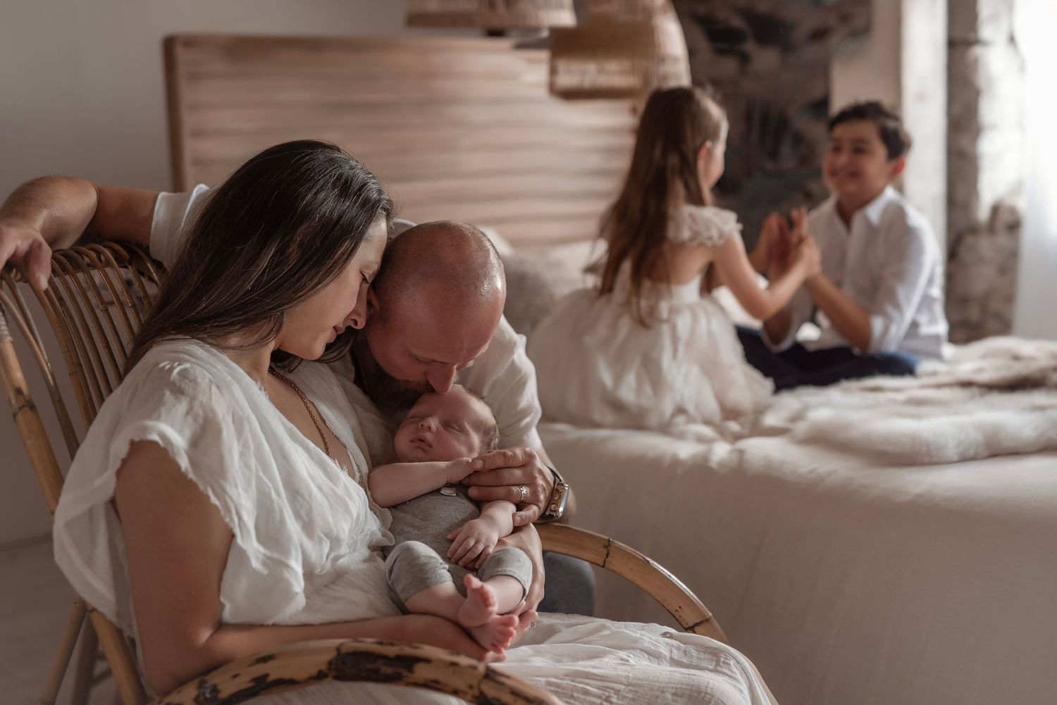 Mother and father in white outfits cuddling newborn baby in rocking chair with children nearby.