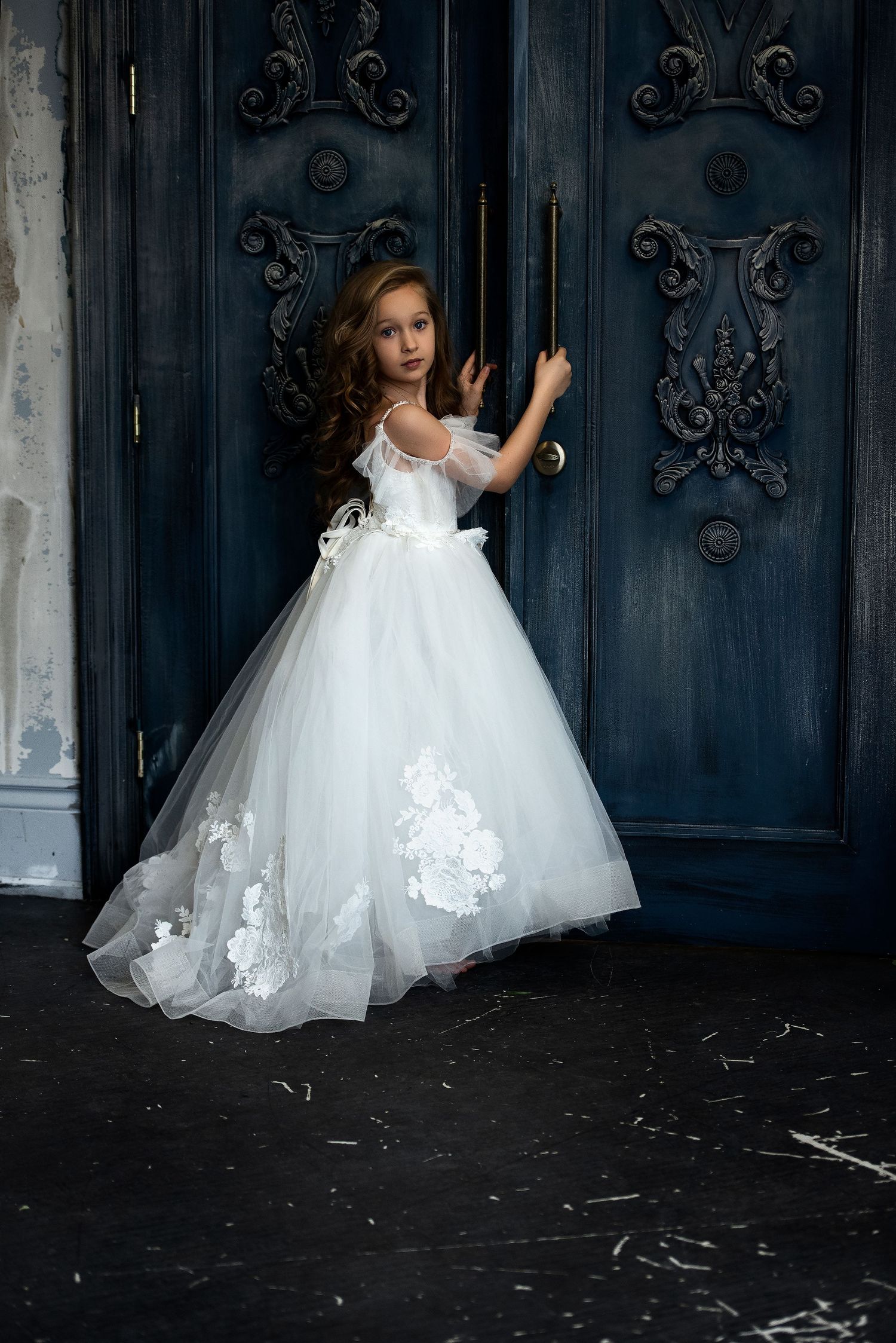 A young girl in a white floral tulle ball gown stands before ornate blue doors.