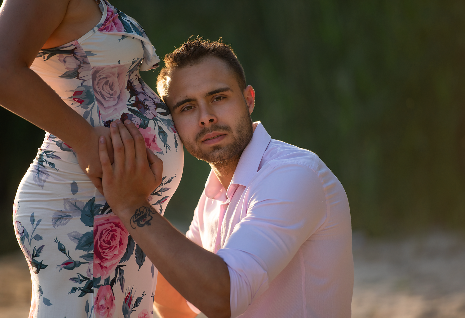 Man in pink shirt kneeling beside pregnant woman in floral dress, gently touching her baby bump outdoors.