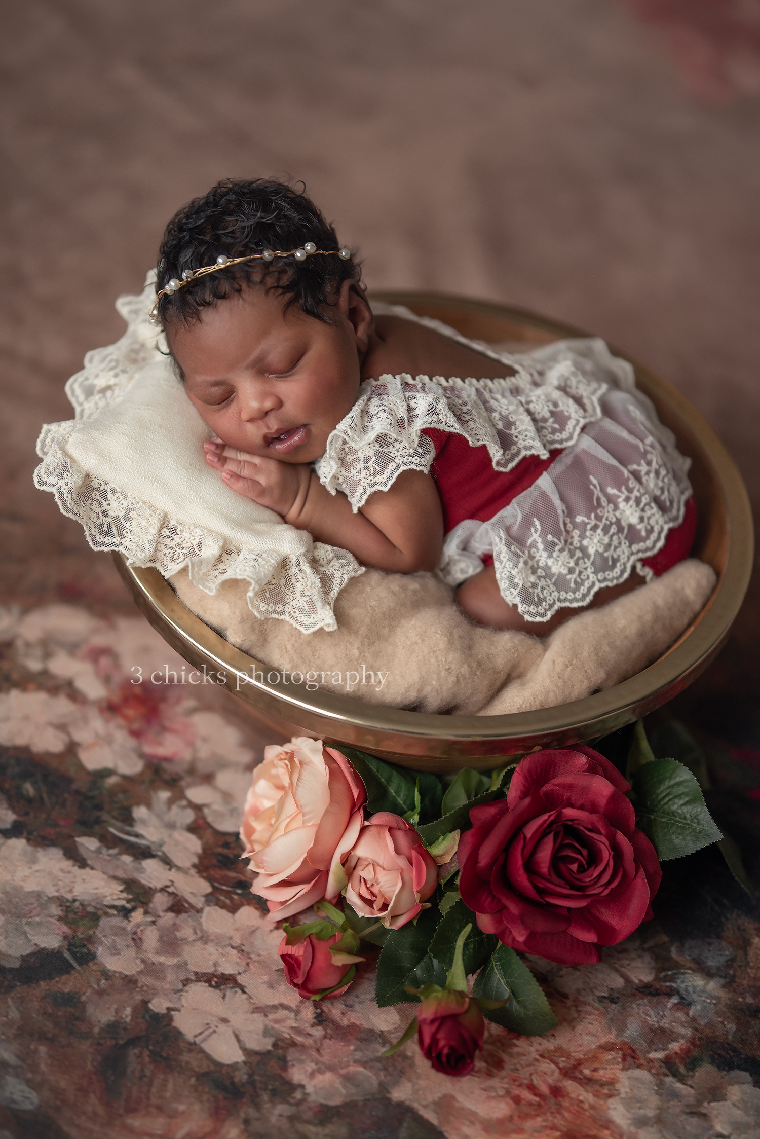 Newborn baby girl sleeping in gold bowl wearing red dress with lace overlay and floral headband surrounded by roses.