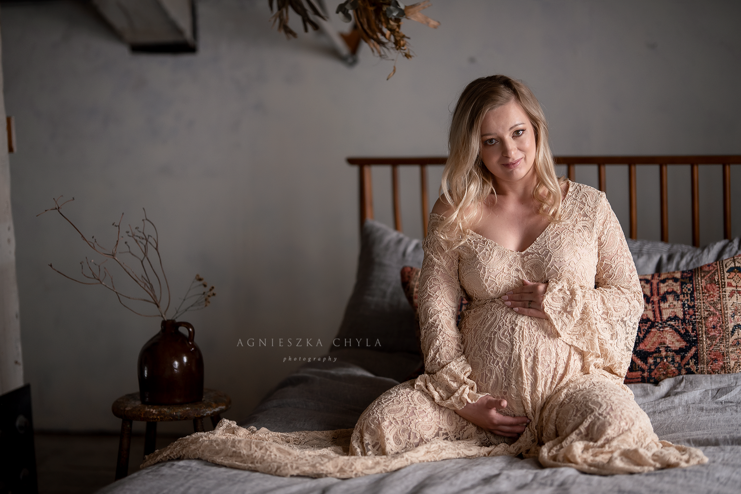 Pregnant woman in lace maternity gown sitting on bed, cradling baby bump in soft natural light.