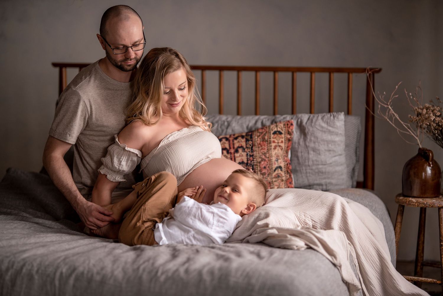 Expectant couple sits on bed as young child rests against pregnant mothers bare belly in cozy bedroom.